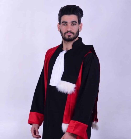 Un homme avec une barbe et des cheveux coiffés porte la « Tenue Doctorale de cérémonie », qui se compose d'une robe noire avec des accents rouges et une bordure en fourrure blanche. Il porte également un vêtement blanc semblable à une écharpe autour du cou. Il se tient debout sur un fond uni de couleur claire.