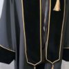 Une robe de cérémonie Toge Premium-Velours en velours noir avec bordure et pompons dorés est présentée suspendue sur un fond blanc. La robe de remise des diplômes comprend une étole de cérémonie avec bordure et pompons dorés assortis aux deux extrémités, conçue pour les cérémonies de remise de diplômes universitaires.