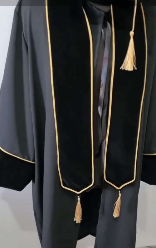 Une robe de cérémonie Toge Premium-Velours en velours noir avec bordure et pompons dorés est présentée suspendue sur un fond blanc. La robe de remise des diplômes comprend une étole de cérémonie avec bordure et pompons dorés assortis aux deux extrémités, conçue pour les cérémonies de remise de diplômes universitaires.